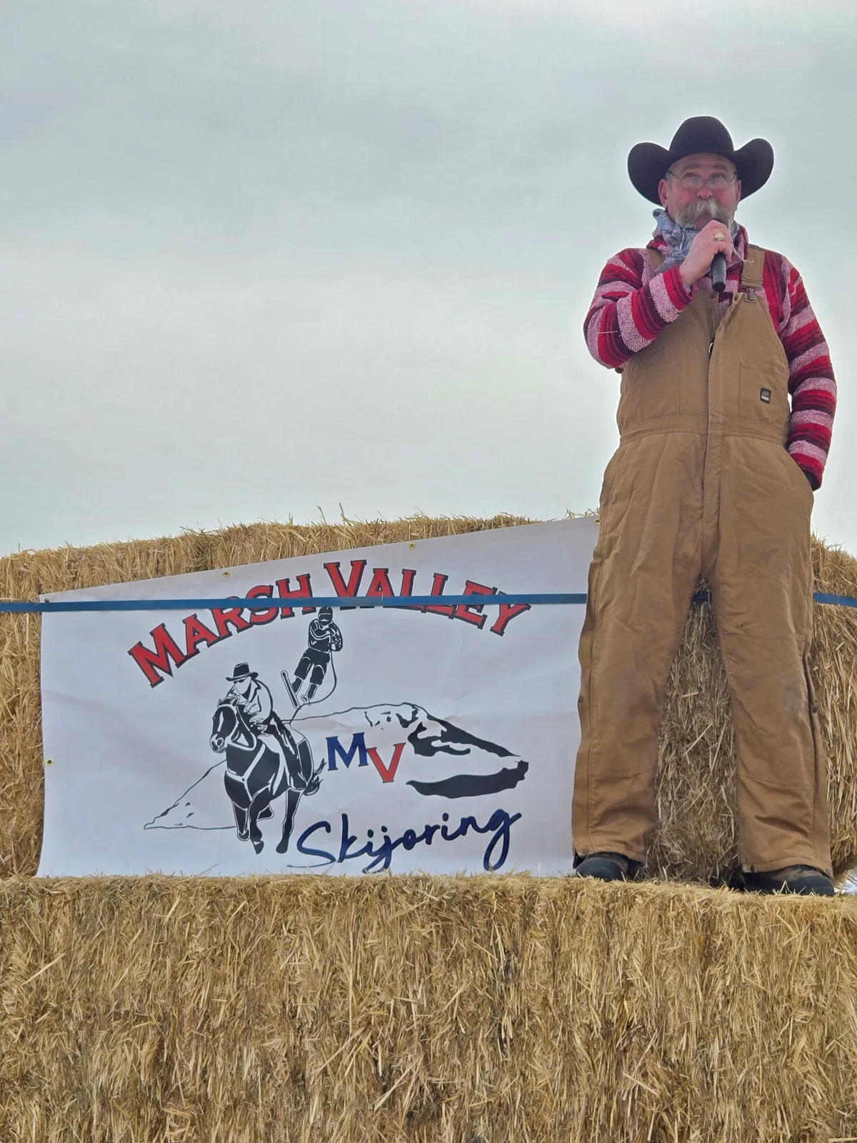Kevin Capson holding a microphone beside the Marsh Valley Skijoring sign