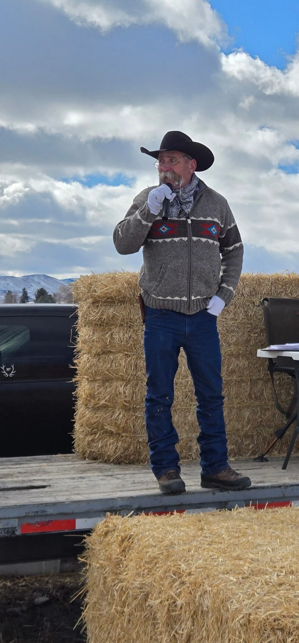 Kevin speaking into a microphone outdoors, wearing a gray Western sweater and black cowboy hat beside hay bales