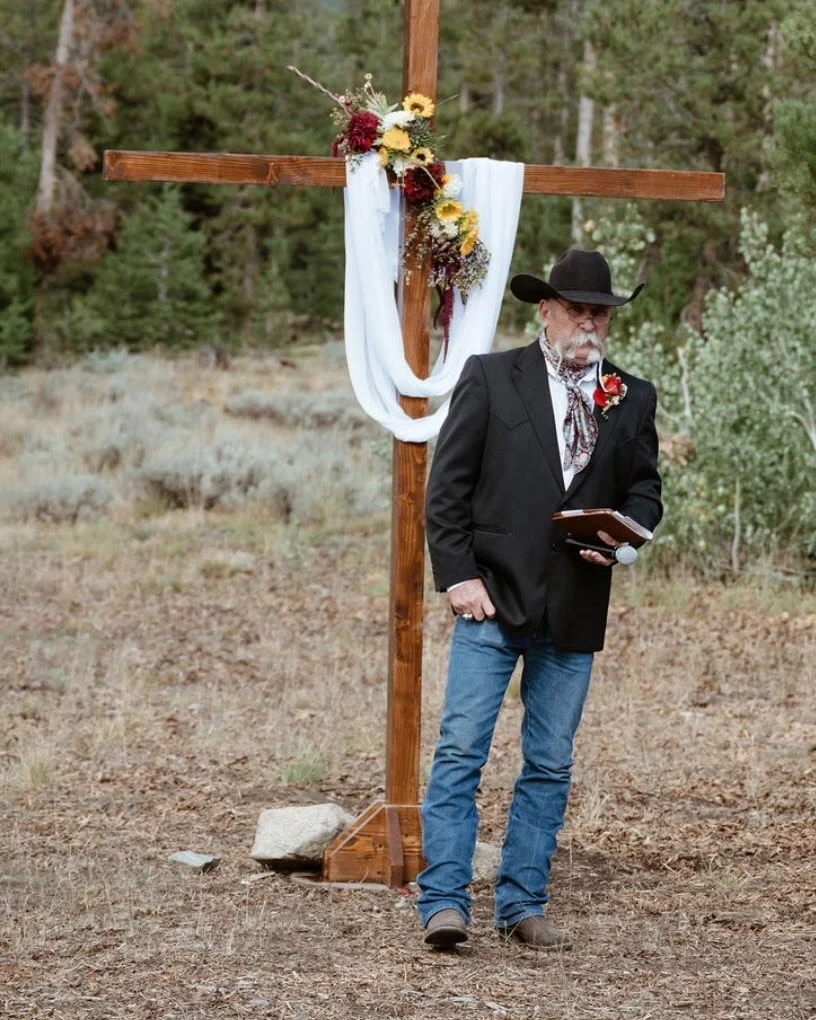 Kevin holding a microphone and ceremony book beside an outdoor wedding arch